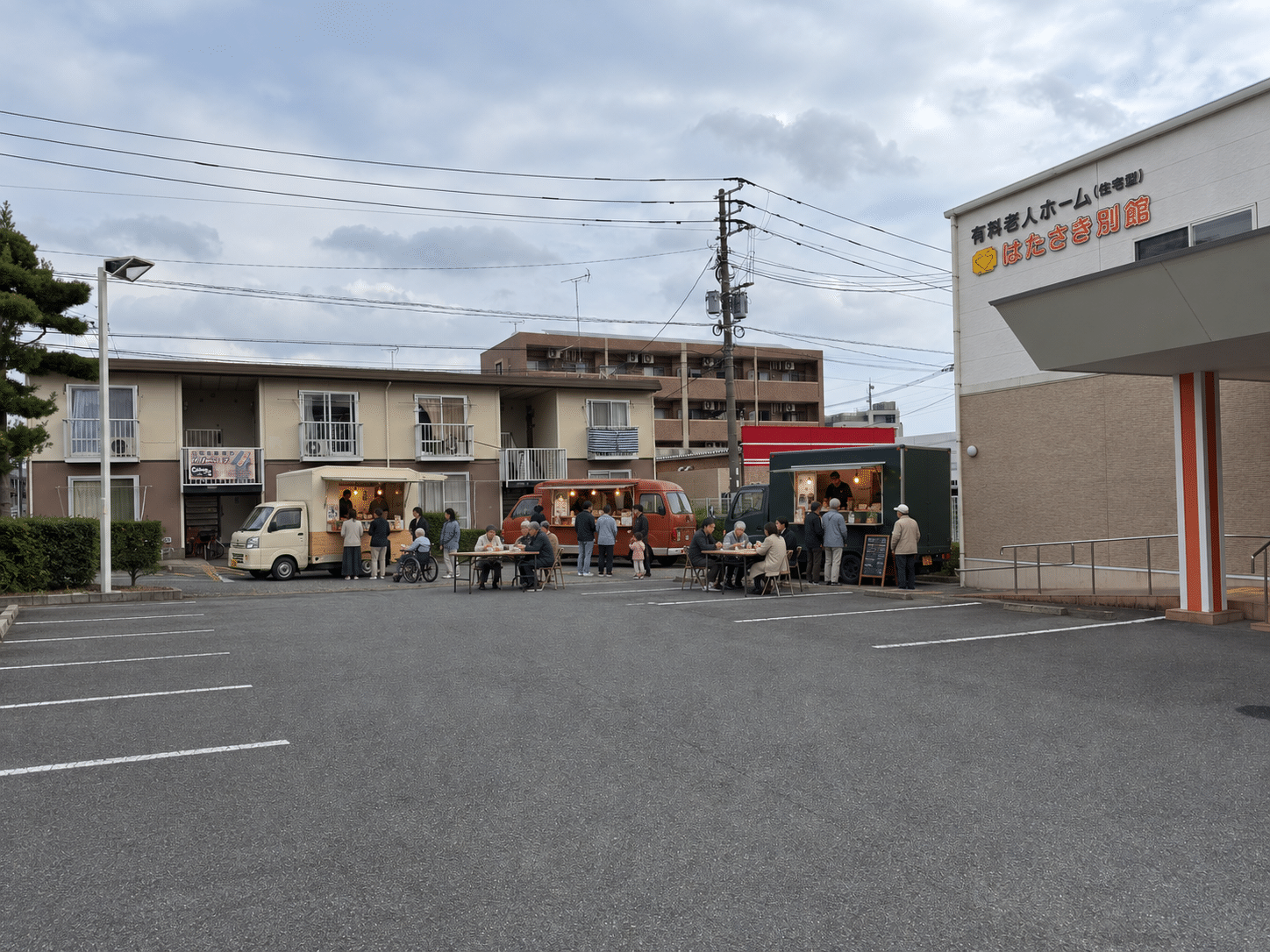 駐車場の奥に3台のキッチンカーを浅い弧状に配置し、近くの長テーブル2台で食事を楽しむイメージ。左側と手前は駐車スペースとして空けています。